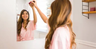 Mujer joven en bata rosa dibujando un corazón en el espejo con pintalabios, sonriendo frente al reflejo