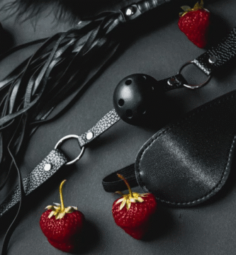Juguetes BDSM como látigo, bola de silencio y antifaz, junto a fresas, sobre fondo oscuro.