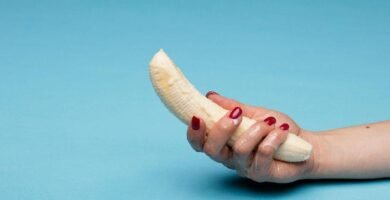 Mano sosteniendo un plátano pelado sobre fondo azul, representación simbólica de la masturbación y el placer masculino.