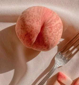 Melocotón entero y trozos con tenedor sobre tela clara, representación artística del erotismo y el placer.